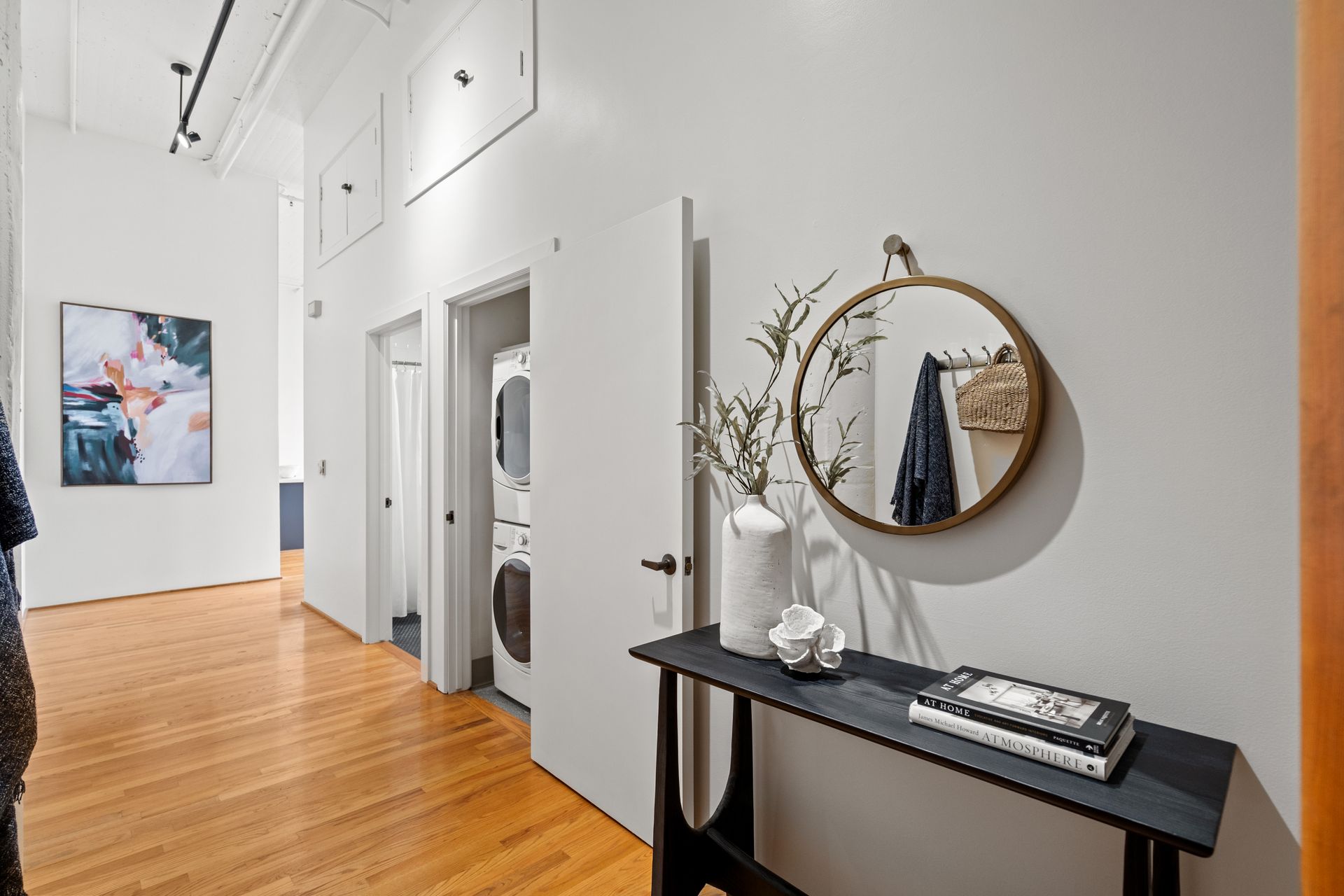 Hallway with hardwood floors, off-white walls, mirror, and decorative table. Artwork and washing machine visible.
