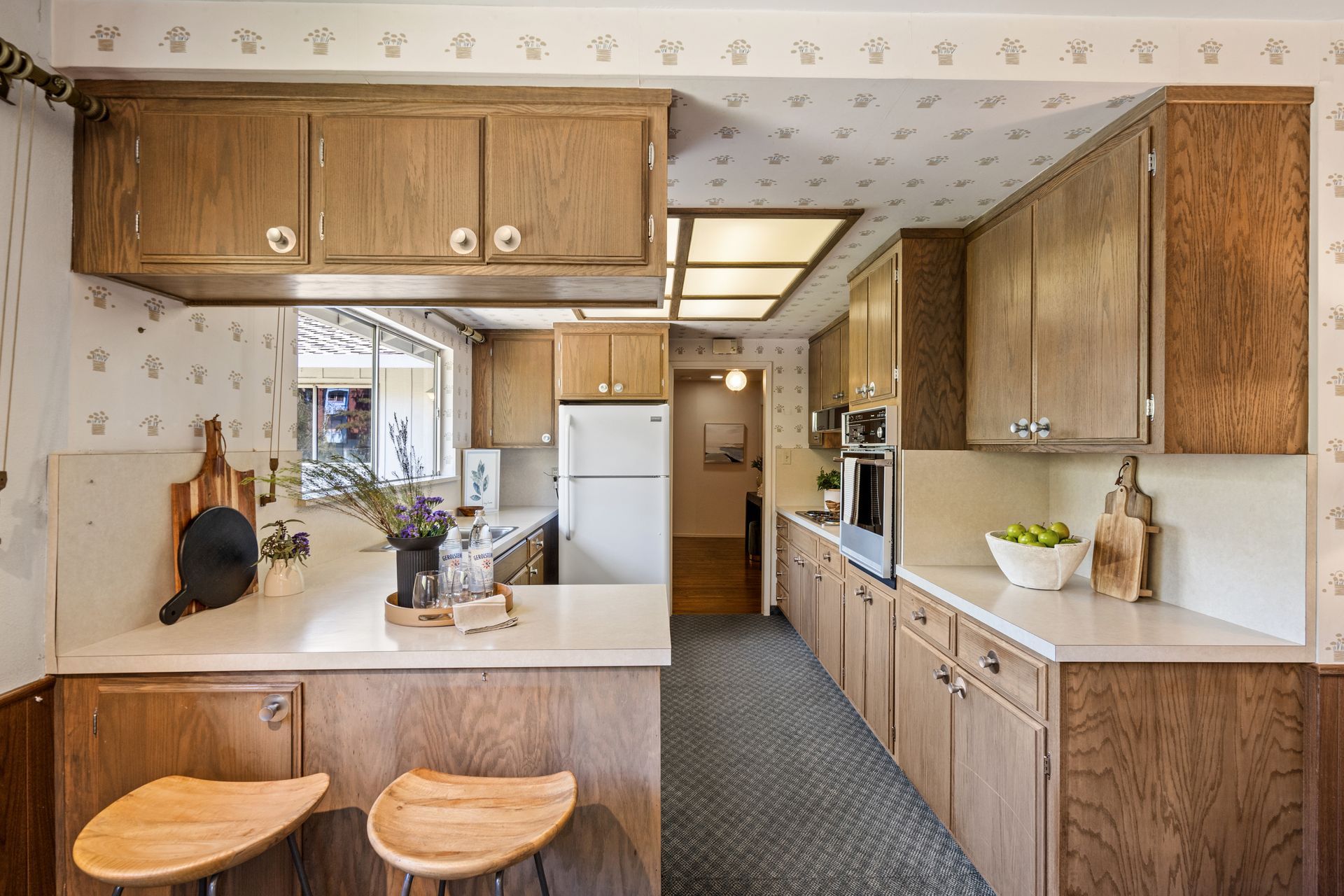 Kitchen with wooden cabinets, white countertops, and a breakfast bar with two stools. The kitchen has patterned wallpaper and a window.