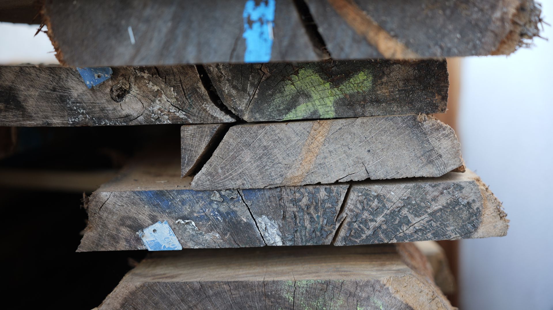Gestapelte, grob zugeschnittene Holzplanken mit dunklen Kanten und blauen Farbflecken, die in Innenräumen gelagert wurden.