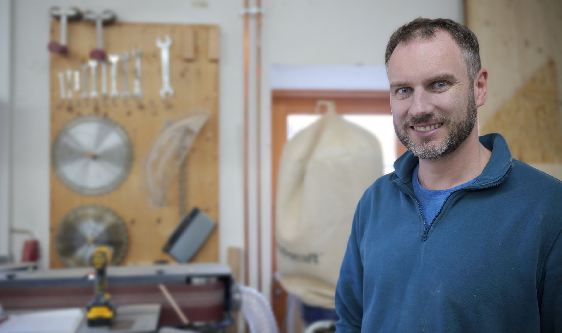 Ein lächelnder Mann in einem blauen Pullover steht in einer Holzwerkstatt, an der Wand hängen Werkzeuge.