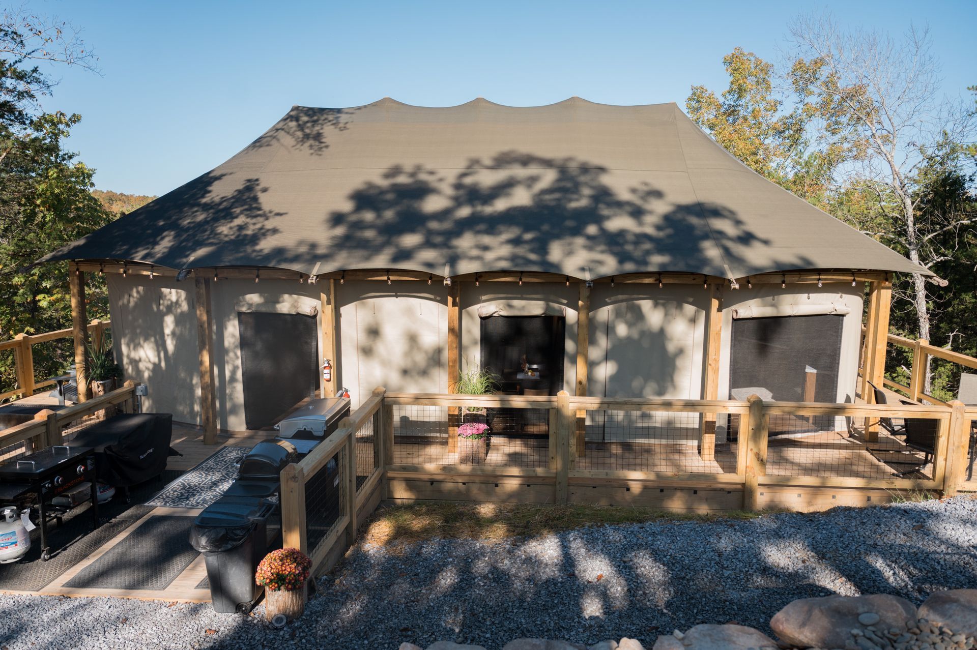 A luxury canvas safari tent structure with a wooden deck and railing, surrounded by trees on a sunny, gravel-covered site.