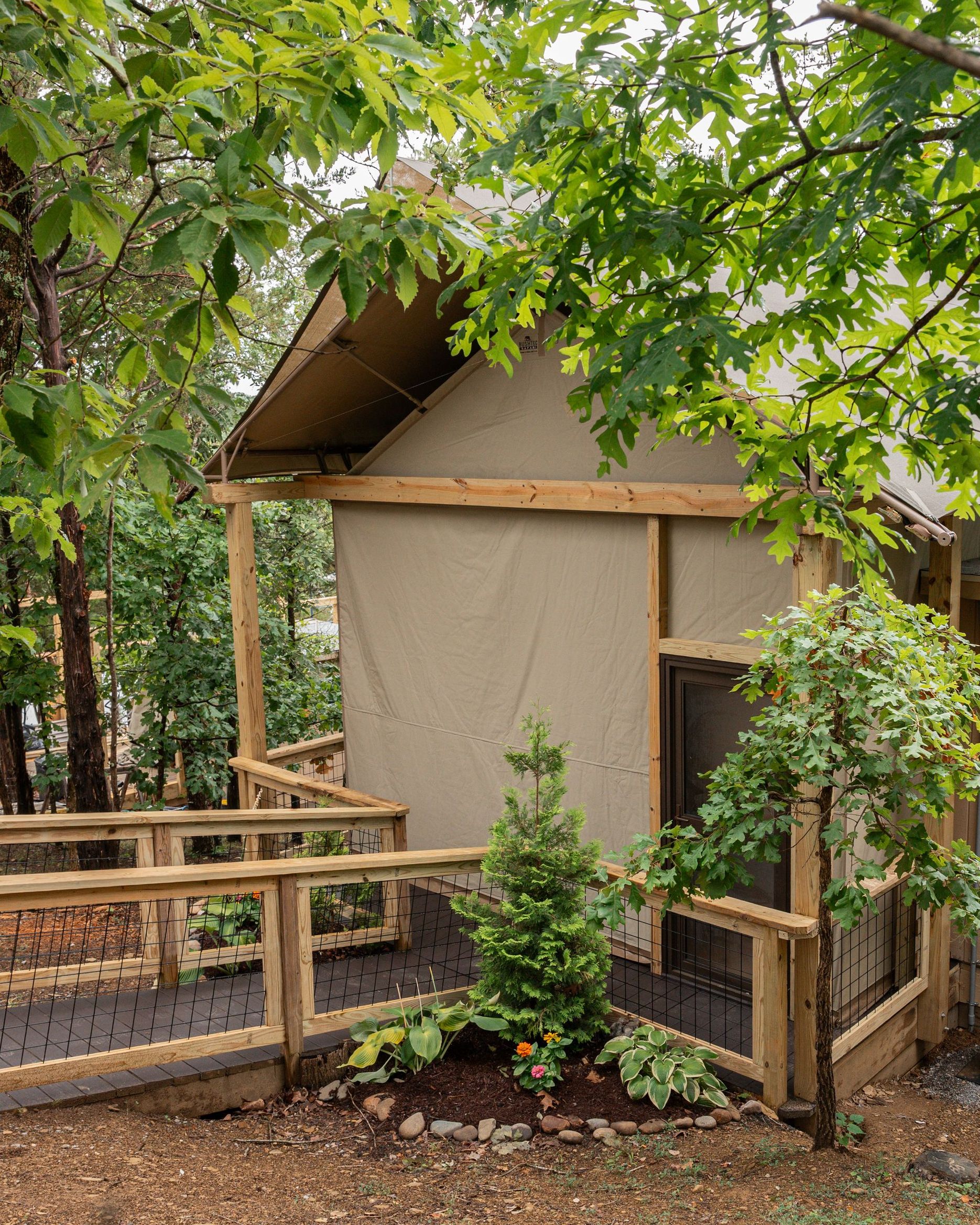 A tan canvas glamping tent with a wooden deck and railing sits nestled among trees in a wooded area.