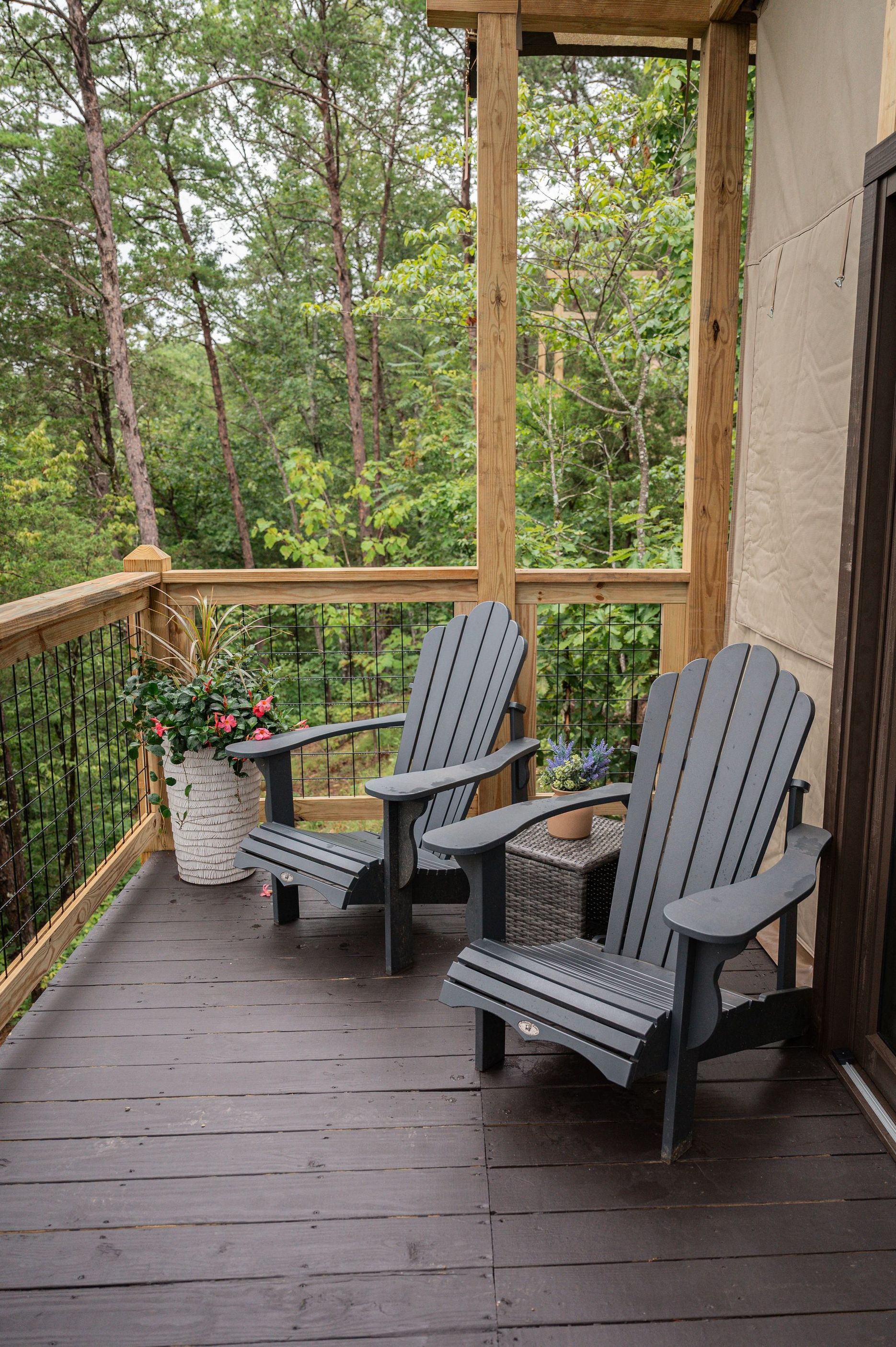 Two gray Adirondack chairs and a small table sit on a wooden balcony overlooking a lush green forest.