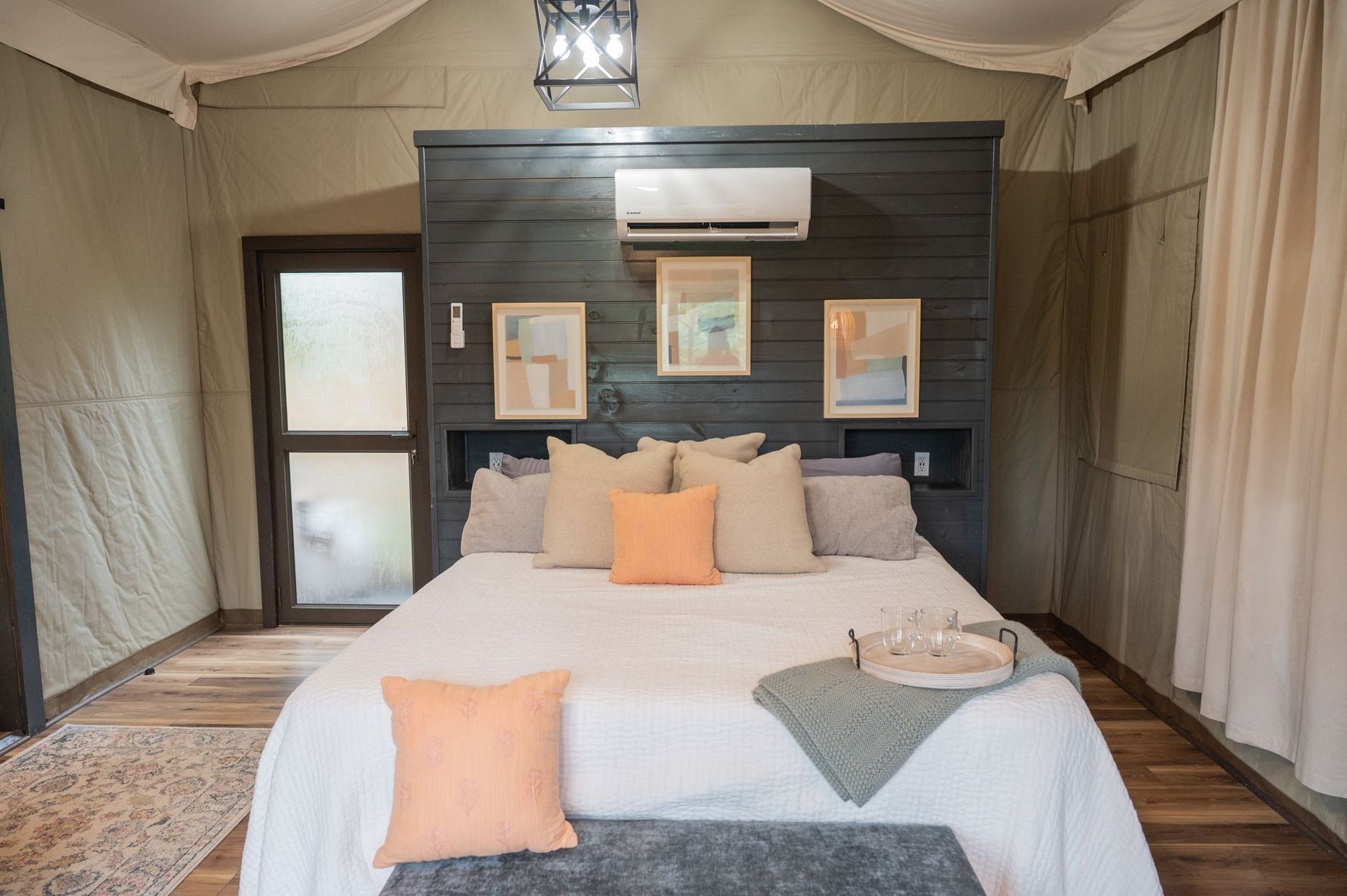 A luxury tented bedroom featuring a bed with neutral pillows and a dark wood paneled headboard with three framed prints.