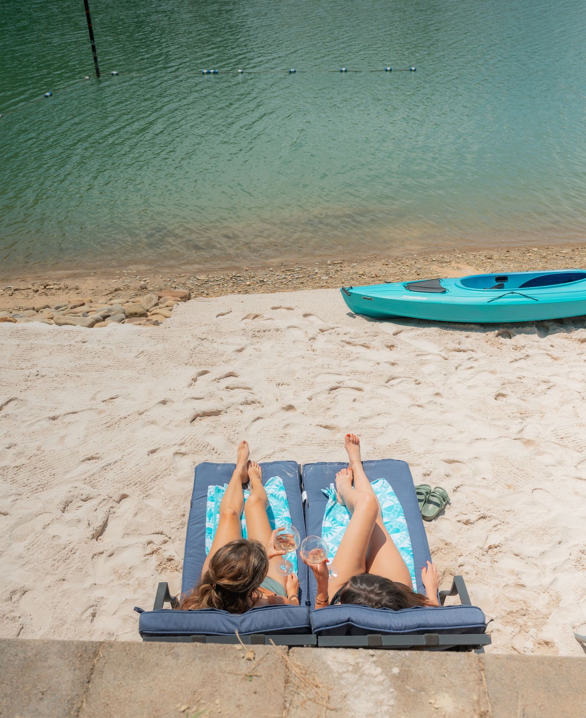 Two people sunbathe on lounge chairs on a sandy beach next to a turquoise kayak by the water.