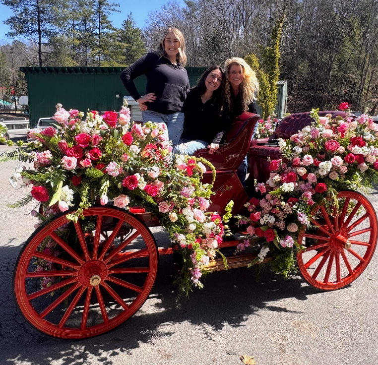 Three people pose behind a vintage, red-wheeled carriage adorned with large, vibrant bouquets of pink and red roses.