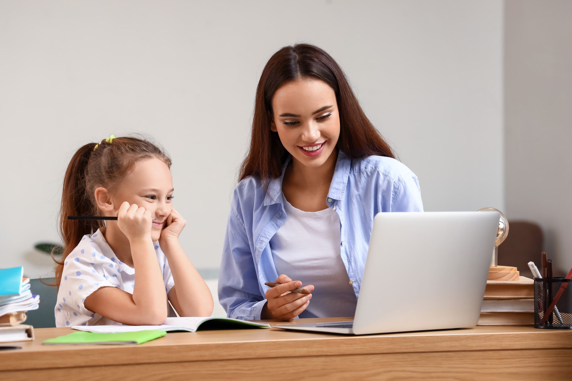 A woman is helping a little girl with her homework while using a laptop computer.