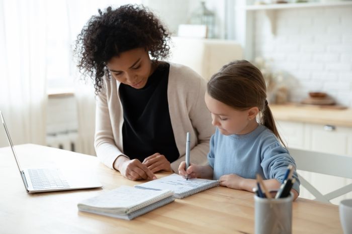 A woman is helping a little girl with her homework while using a laptop computer.