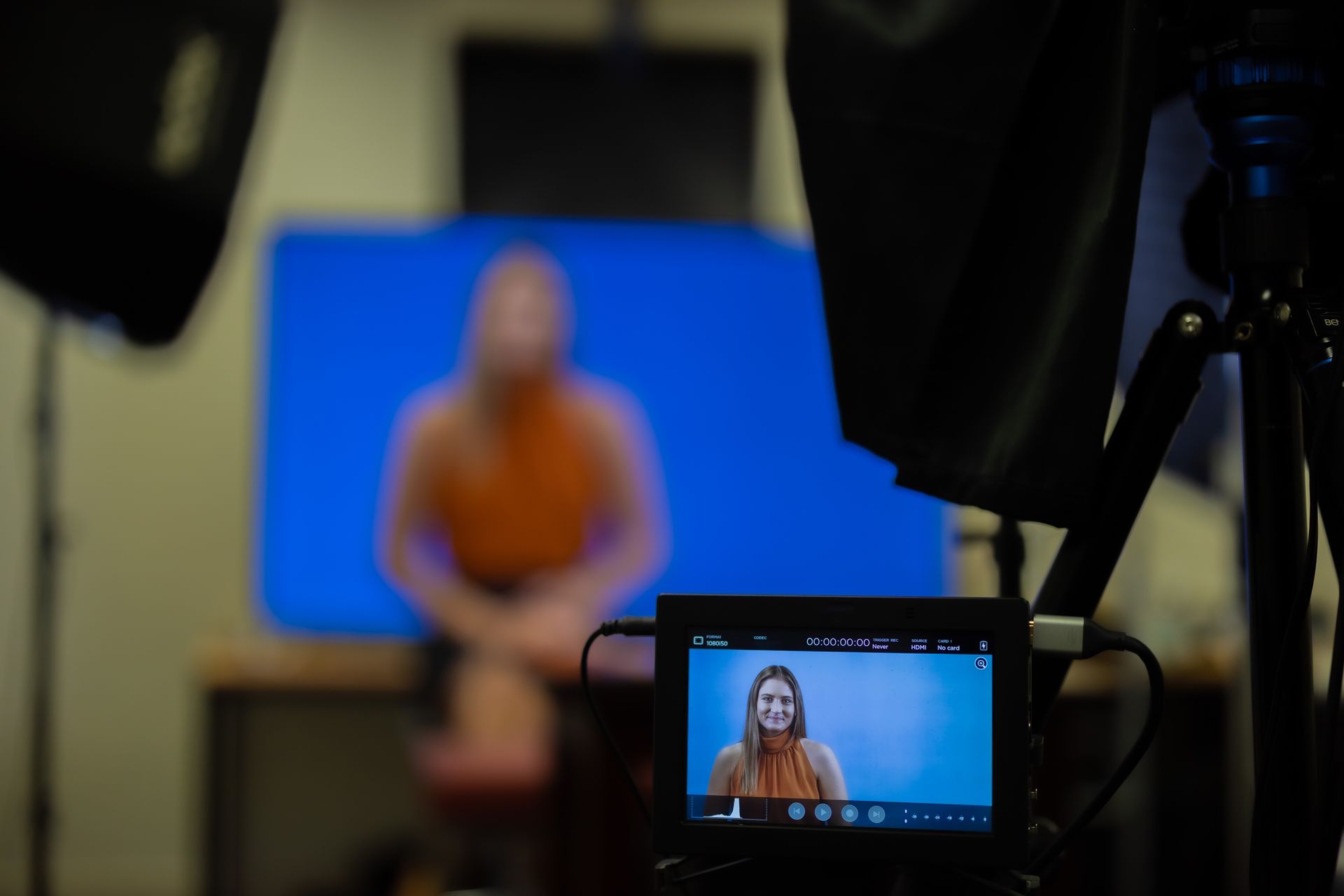 A woman in an orange top sits in front of a blue screen being filmed. A monitor displays her face.