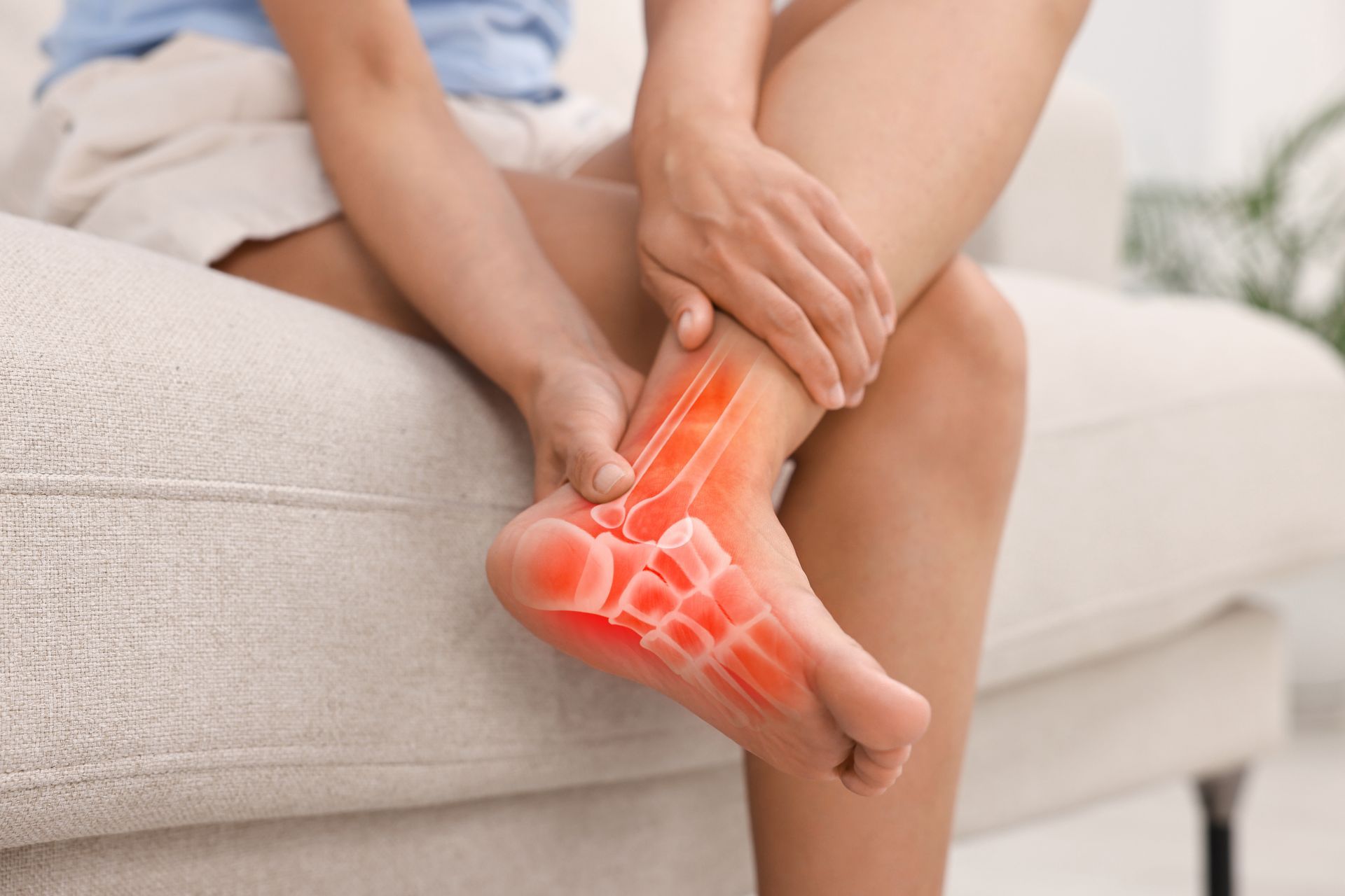 Person holding foot, showing pain in ankle and foot bones highlighted in red. Sitting on a sofa.