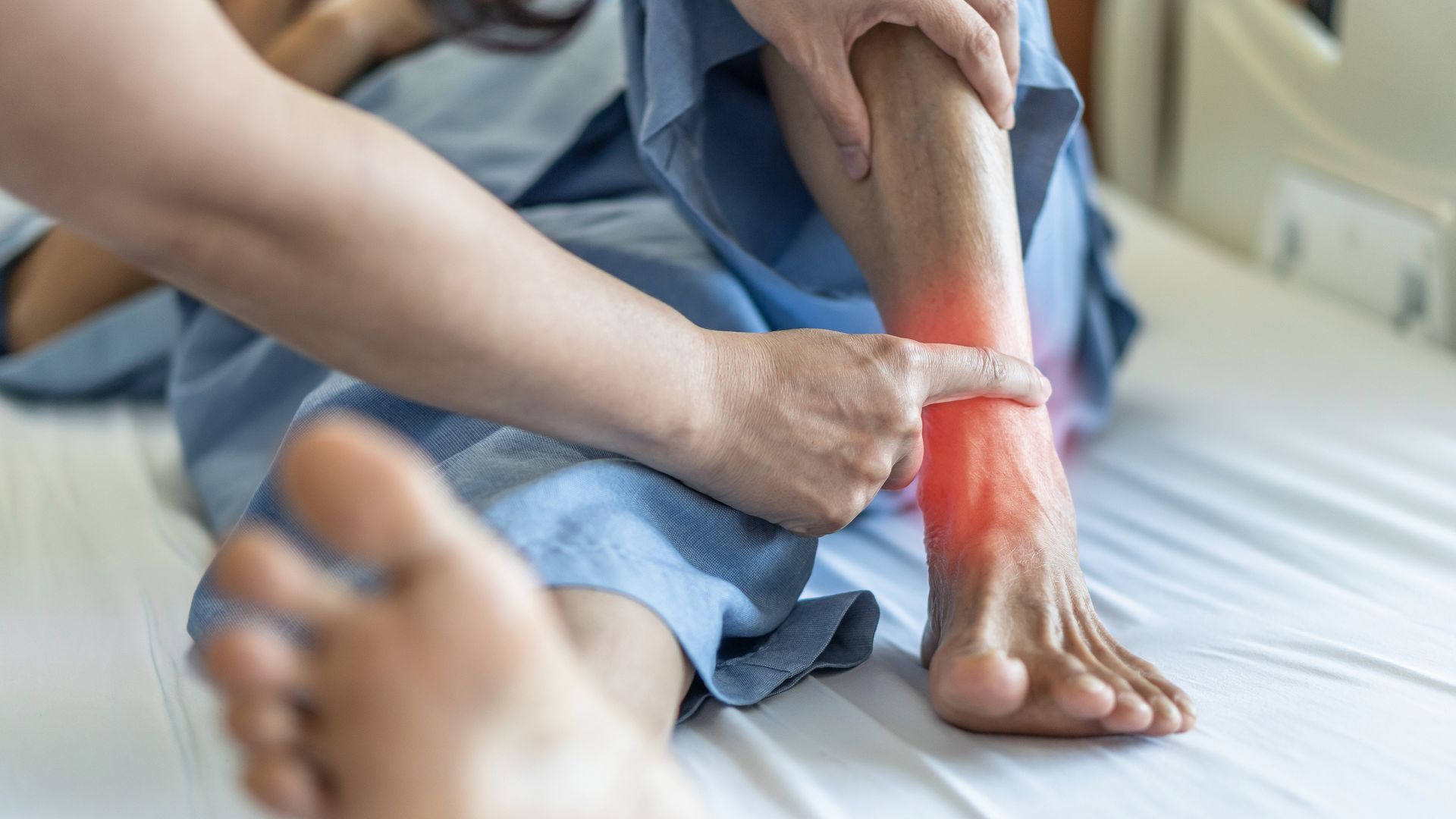 Person's swollen ankle being examined by another person's hand, red area highlighted on leg, indoors, bed.