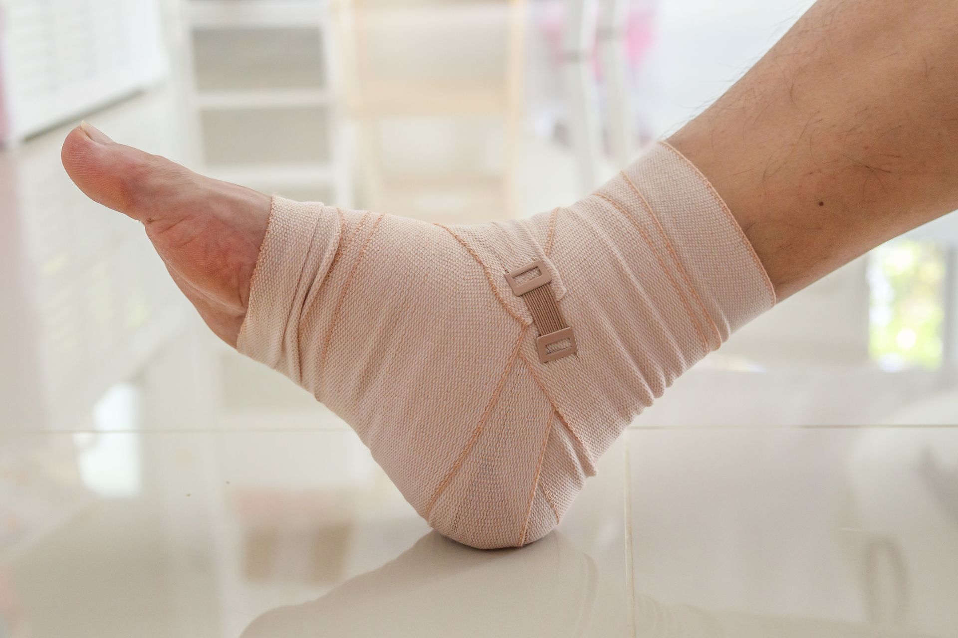 Ankle wrapped in beige medical bandage, resting on a white, shiny floor.