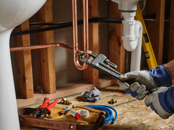 Plumber tightening a copper pipe under a sink with a wrench, tools on the floor in an unfinished room