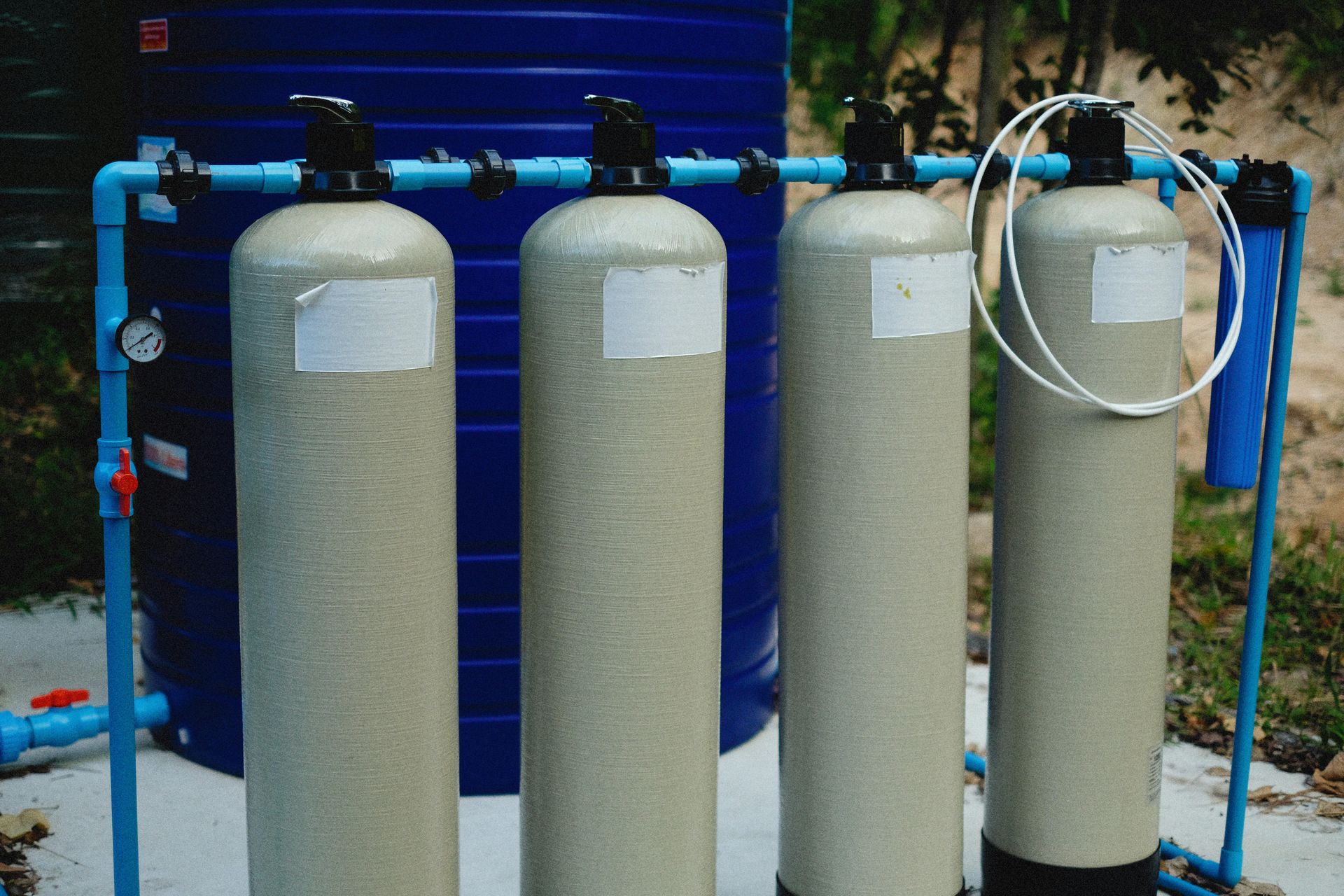Four large gray water filters connected by blue pipes in front of a blue wall.