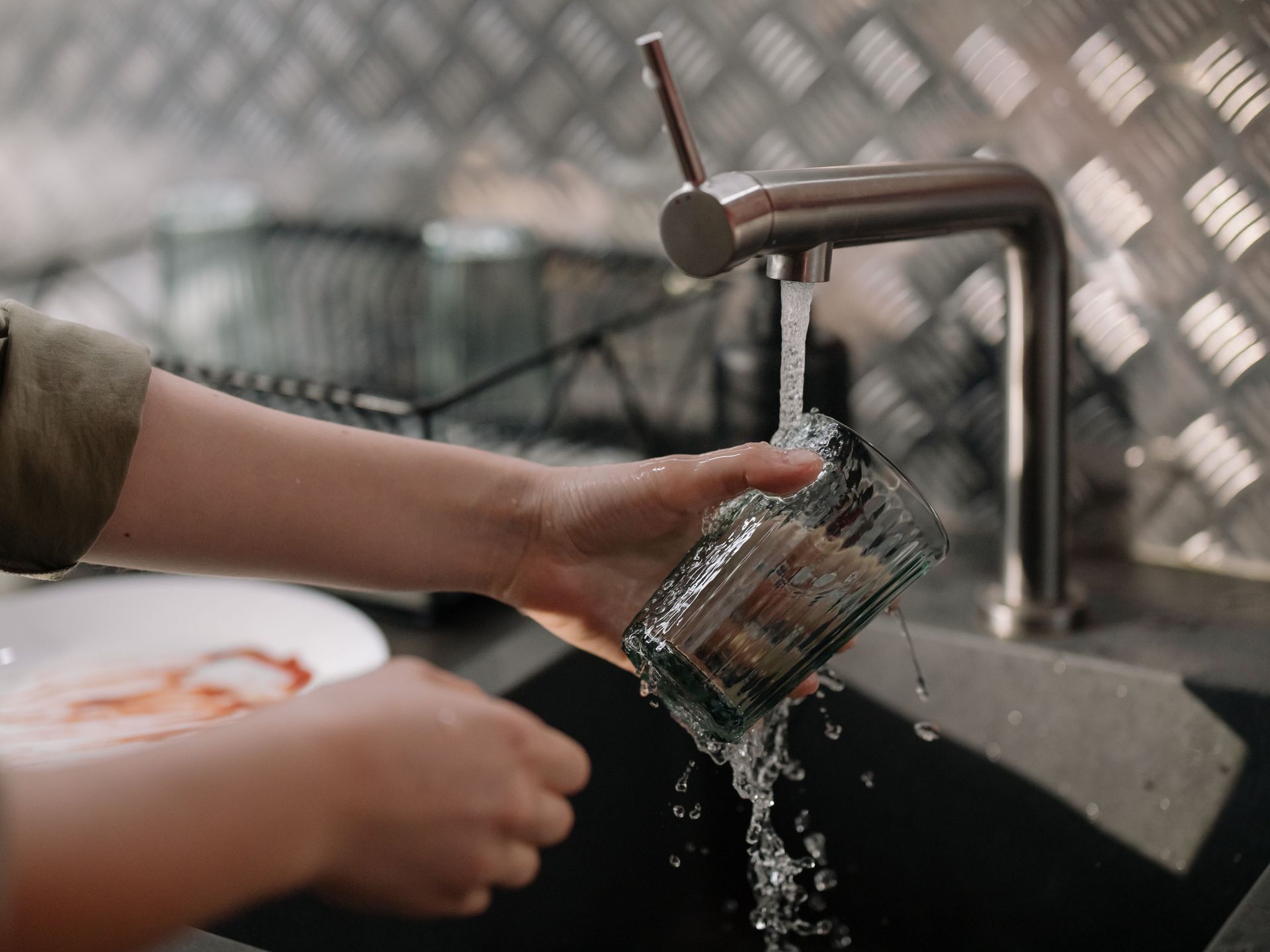 Hand washing a glass under a kitchen faucet, with water splashing into the sink