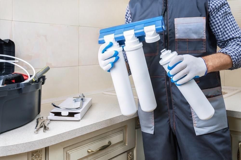 Person holding three white water filter cartridges and a blue filter head in a tiled kitchen or utility room