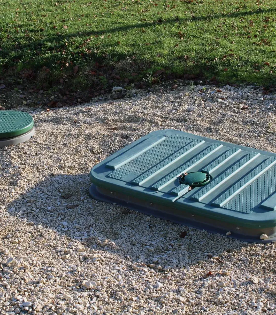 Green utility cover and round lid on gravel beside a grassy area