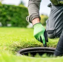 Worker in green gloves using a vacuum hose to clean a lawn drain in a grassy yard