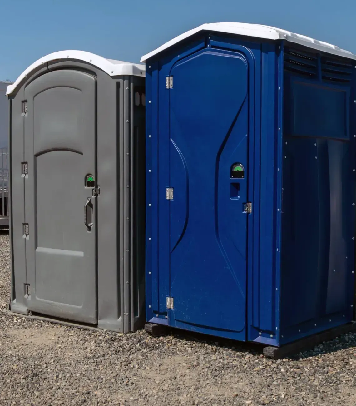 Two portable toilets, one gray and one blue, standing on gravel outdoors under bright sunlight