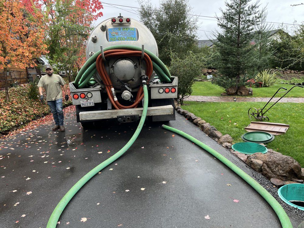 Worker pumping water from a tanker truck into a flooded driveway; hoses run across the wet pavement.