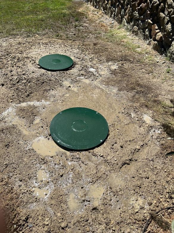 Two green circular utility covers set in bare dirt on a sloped roadside.
