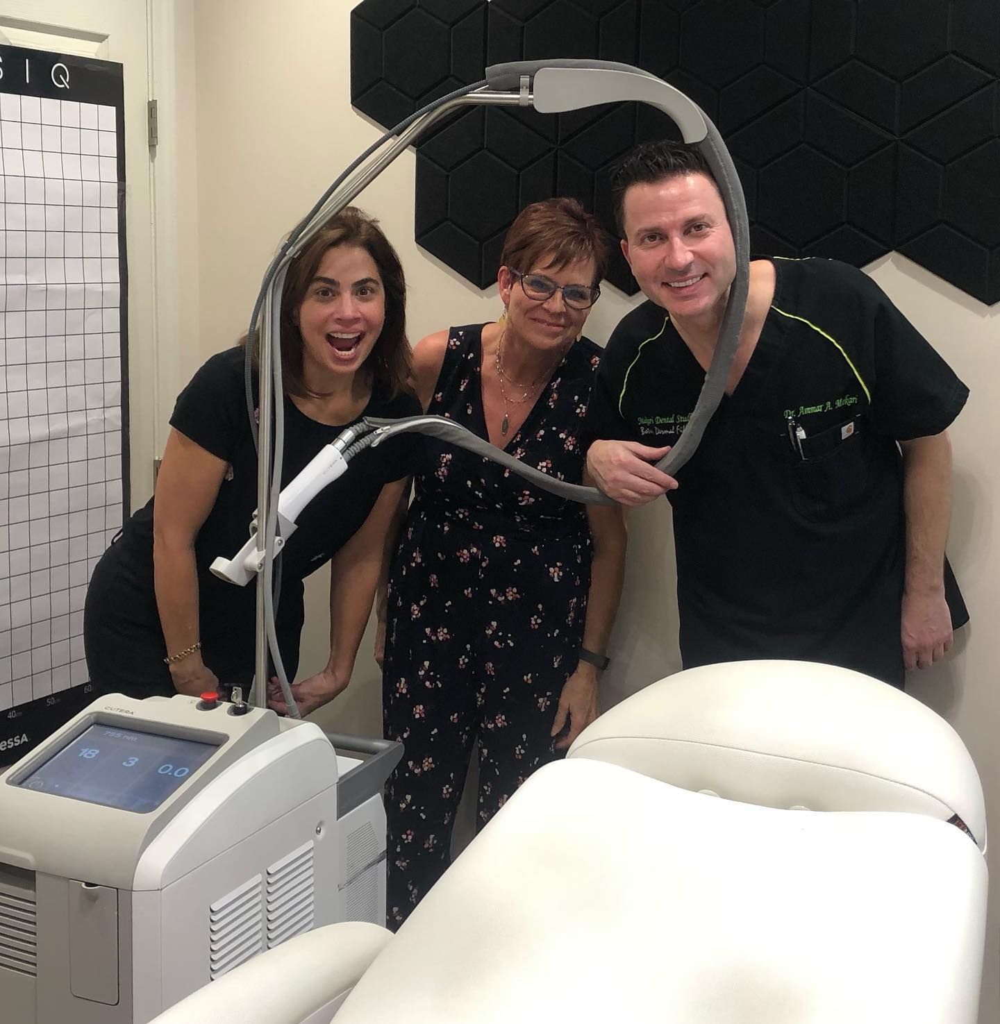 Three people pose with a laser machine in a clinic setting. Smiling, they appear to be staff and patient.