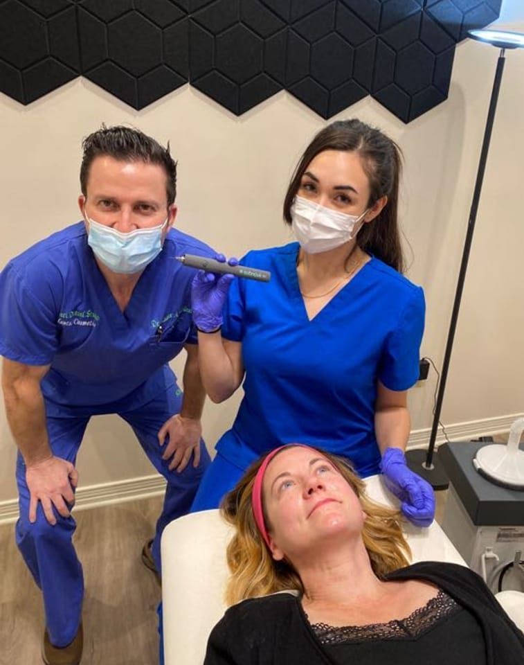 Two medical professionals, one holding a tool, over a person on an exam table; blue scrubs, masks, medical setting.