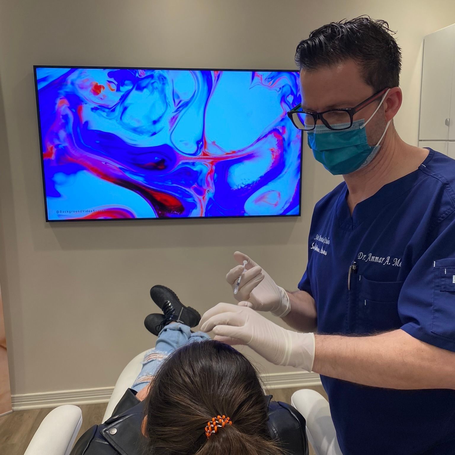 Doctor in scrubs, mask, and gloves performing a procedure on a patient in a medical office.