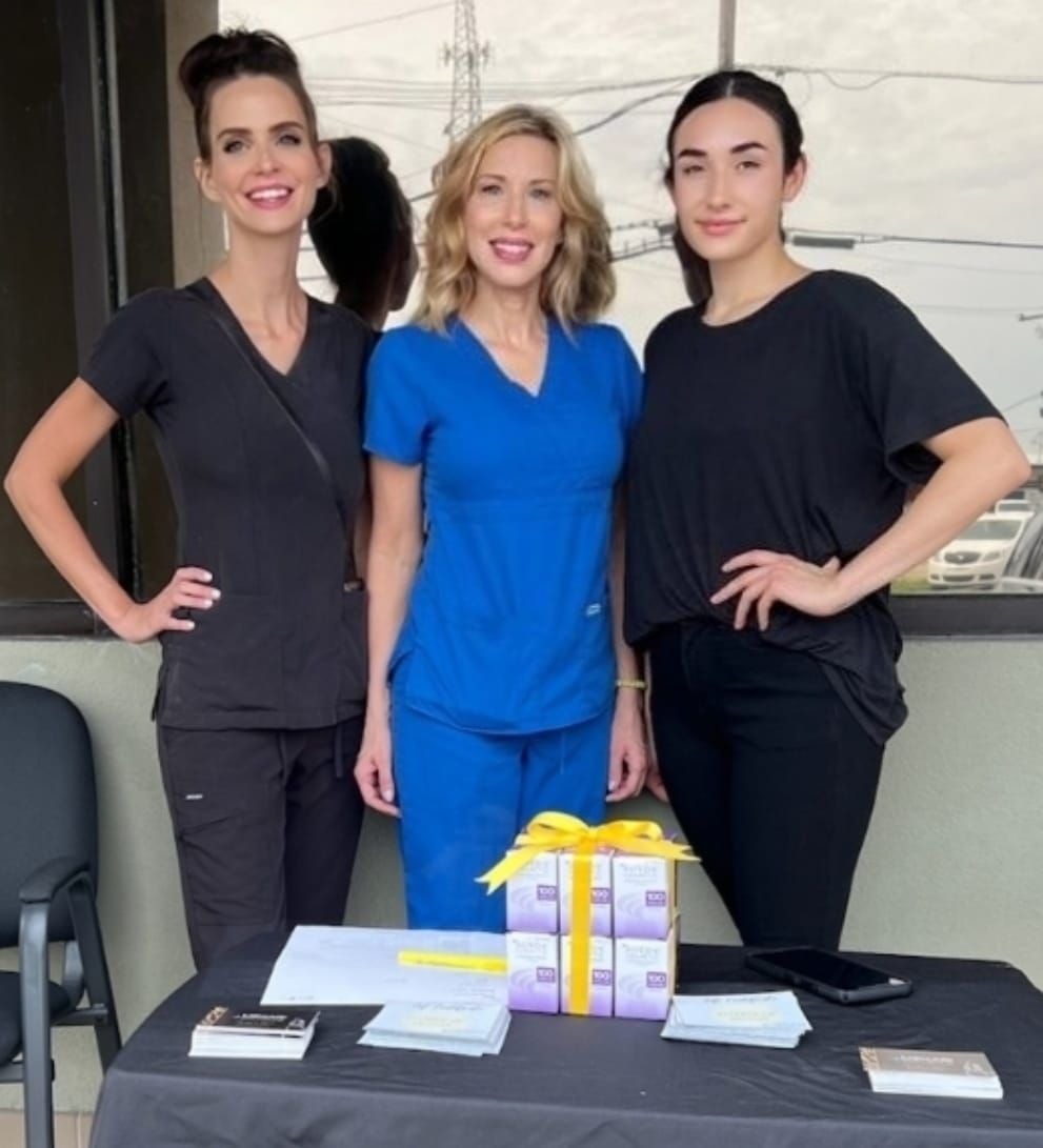 Three women at a table. Two in black, one in blue scrubs. Smiling, hands on hips, near a gift box and papers.