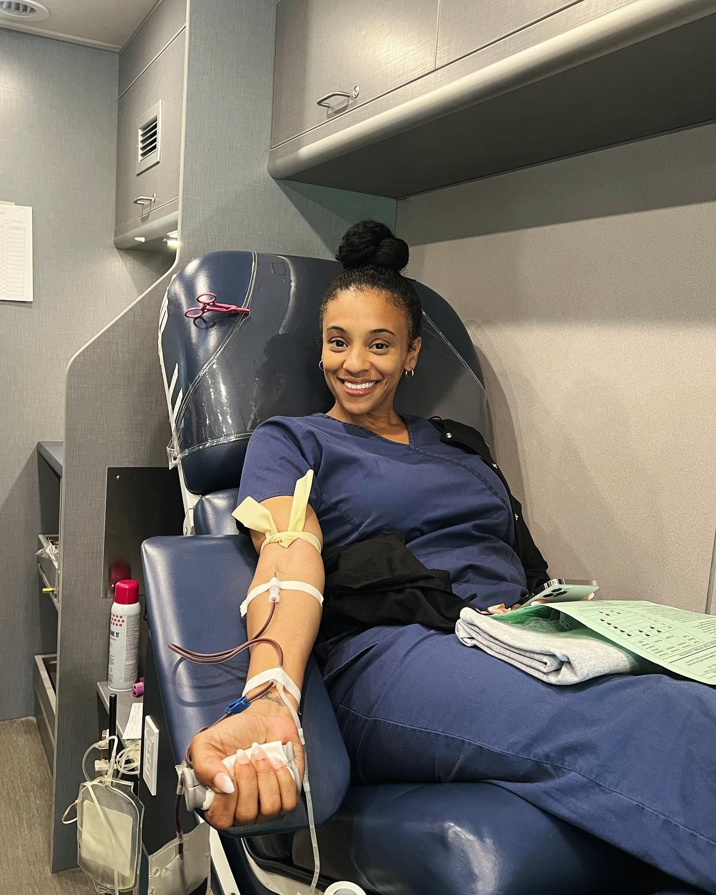 Person in blue scrubs donating blood in a mobile blood donation center; smiling, arm connected to blood bag.