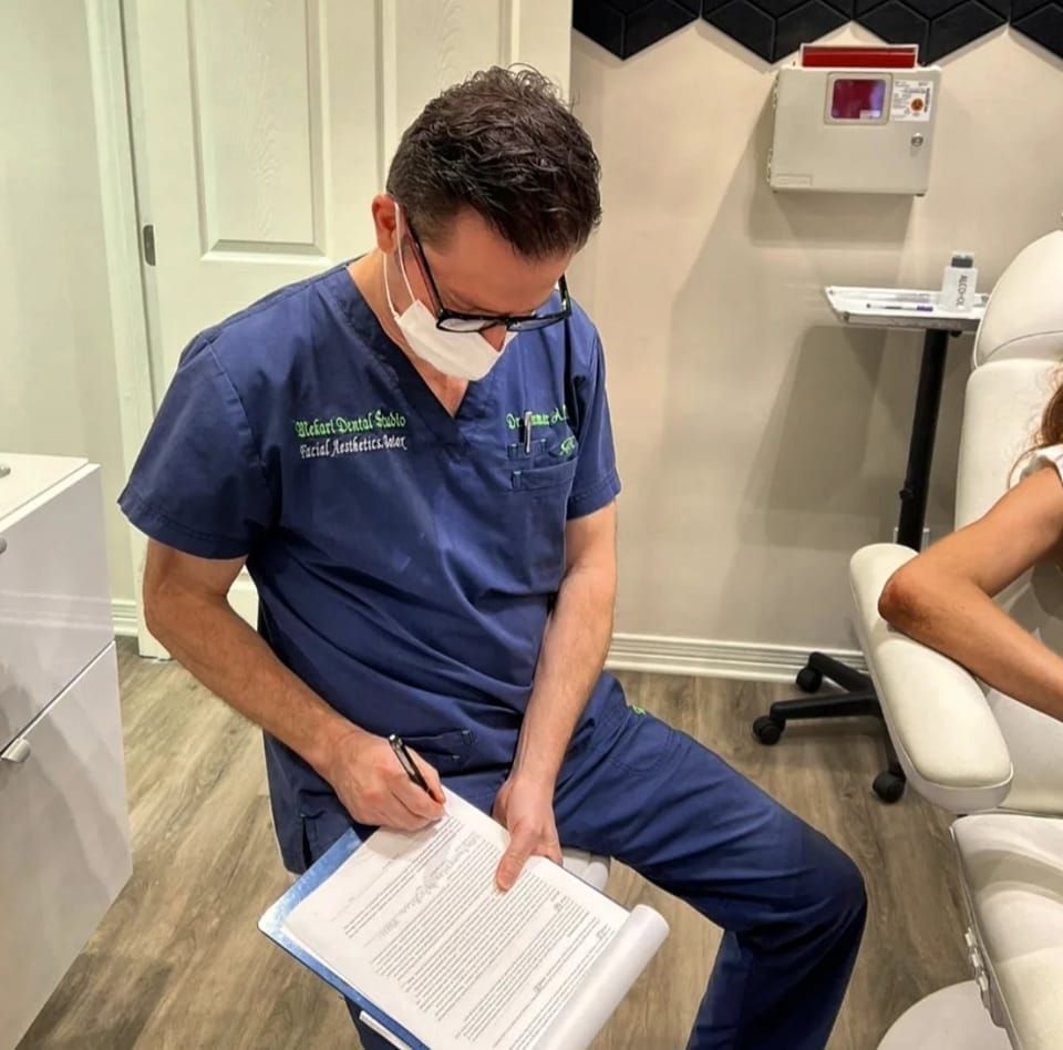 Doctor in blue scrubs, mask, and glasses reviewing paperwork near a patient in a medical office.