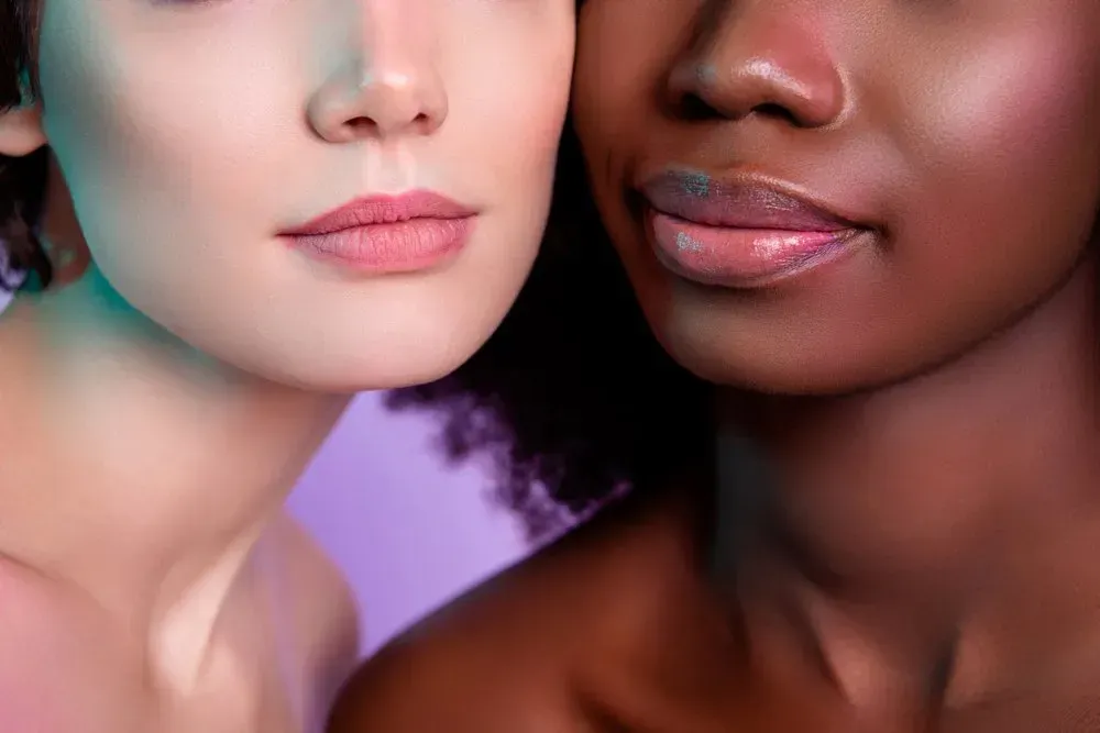 Two people, diverse skin tones, close-up on faces. One fair, one dark, both with glossy lips, and soft lighting.