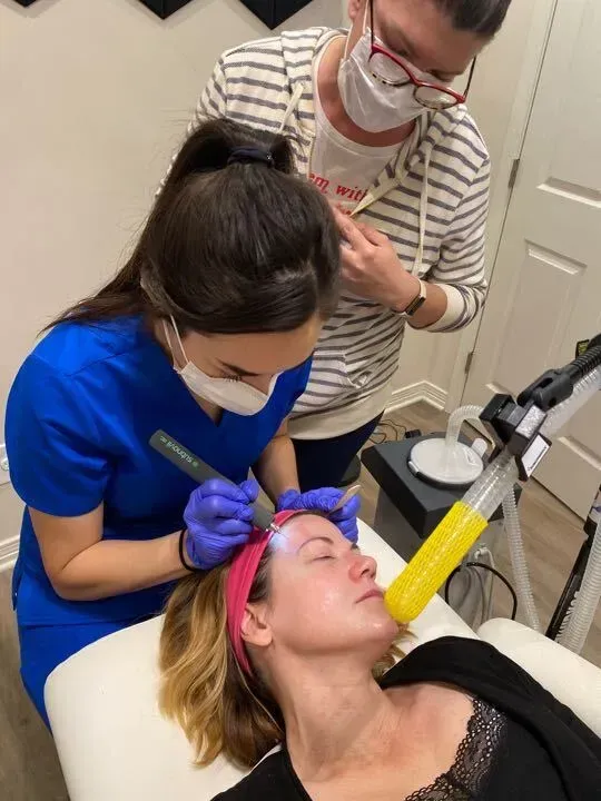 Woman receiving facial treatment in a clinic. Two staff members wearing masks, one holding a device near the patient's face.