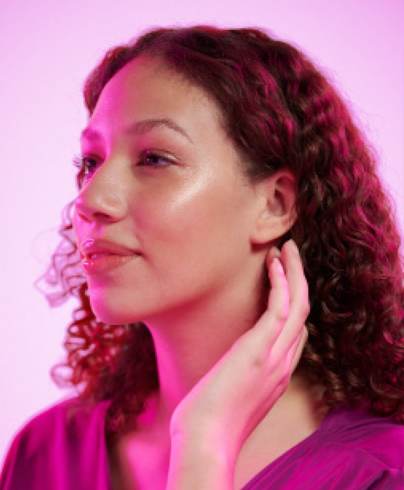 Woman with curly hair touches her neck, lit with pink light.
