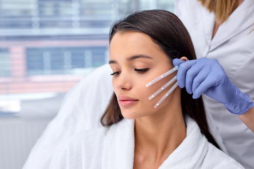 Woman receiving facial treatment, lines drawn on cheek, gloved hand holds needles.