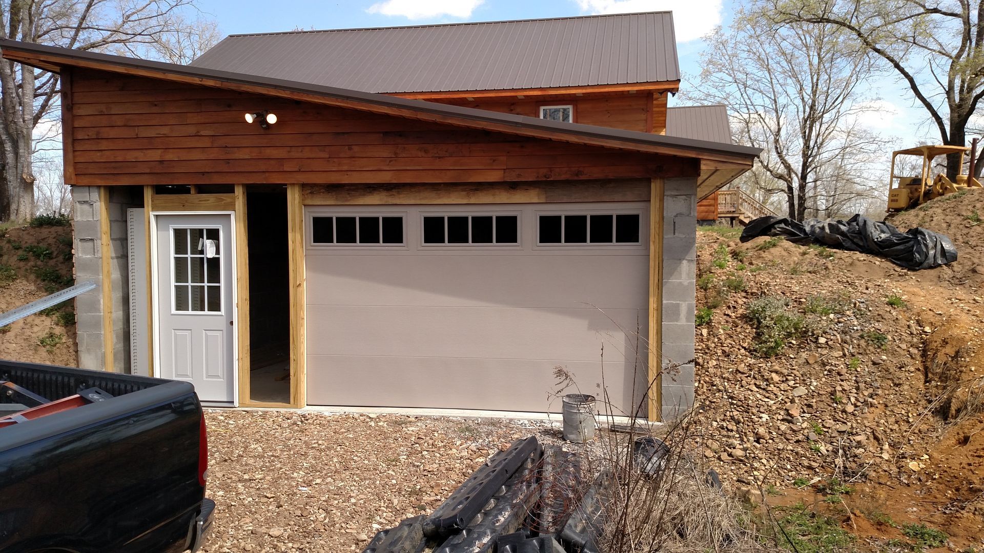 Brown house with attached garage and white door, surrounded by gravel and leaf-covered hillside.