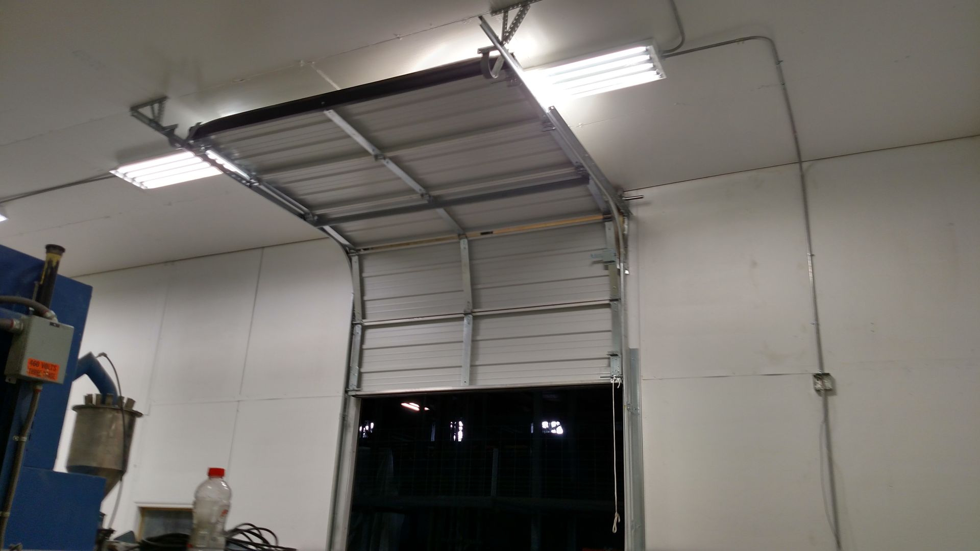 Interior of a warehouse with a partially open roll-up garage door under bright ceiling lights.