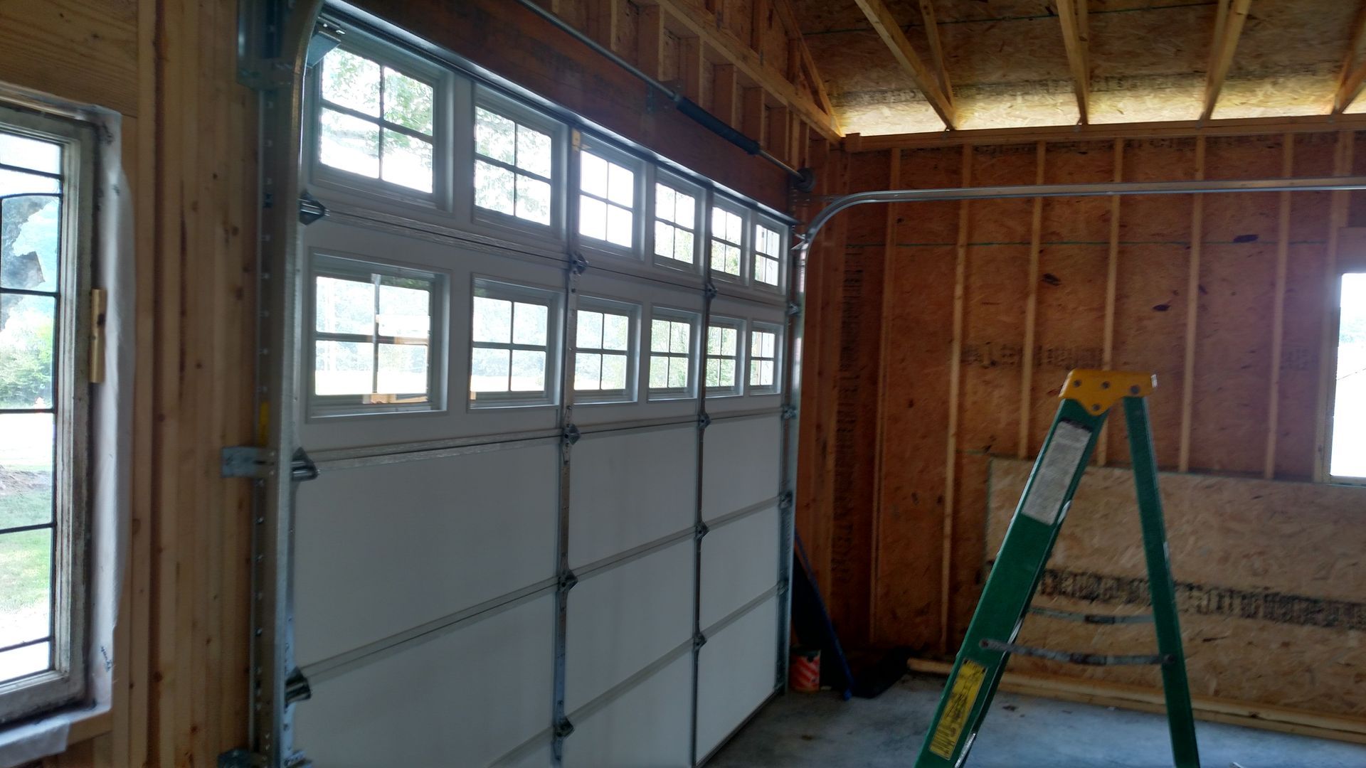 Inside a garage, a white roll-up door, wooden walls, and a green ladder near a window.