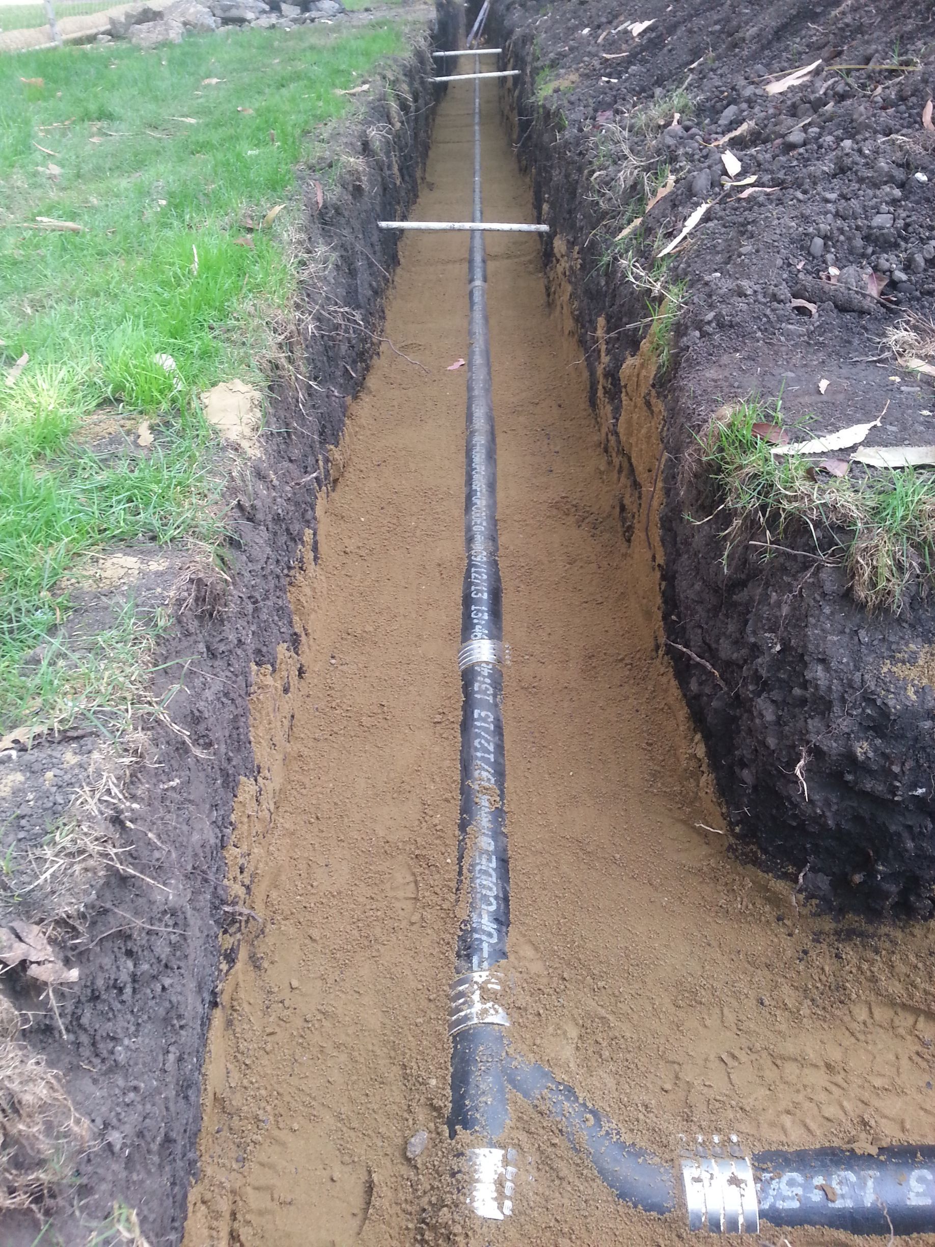 Black pipe laid in a trench, prepared for burial. Dirt and grass surround it.