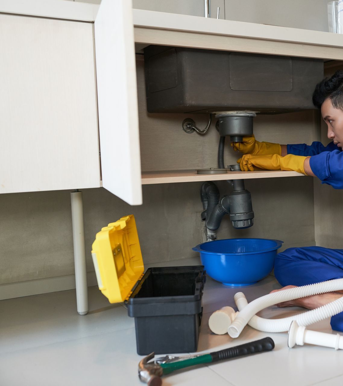 Plumber fixing pipes under a kitchen sink, wearing gloves. Tools and a blue bowl are on the floor.