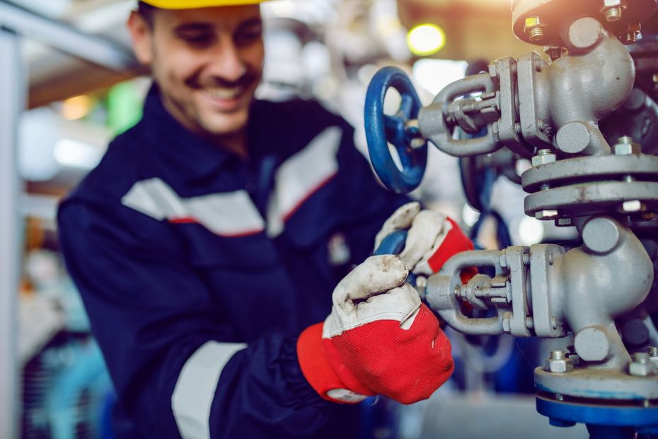 Man in uniform and gloves turning a valve with a smile in a mechanical setting.