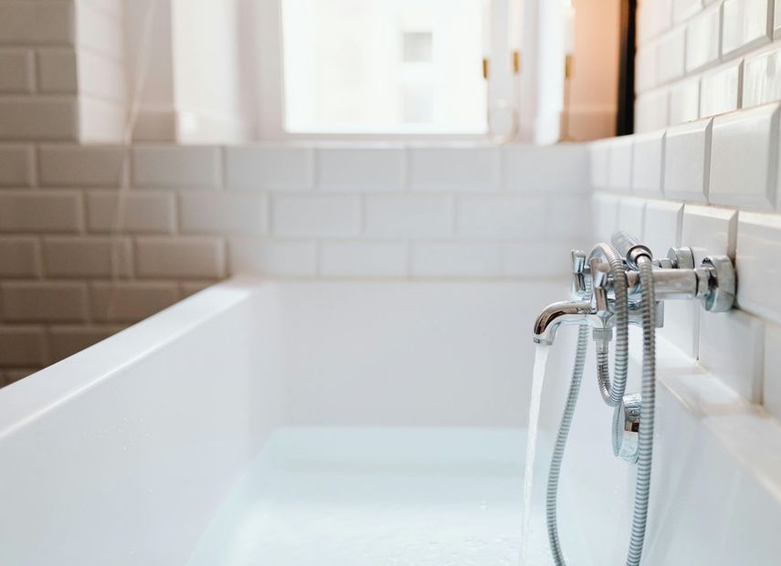 Bathtub filled with water, faucet running; white tile wall, window in background.
