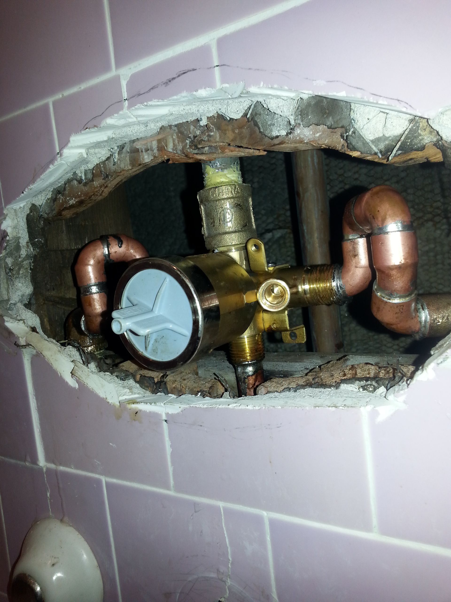 Shower valve assembly with copper pipes exposed in a wall opening; pink tiles.