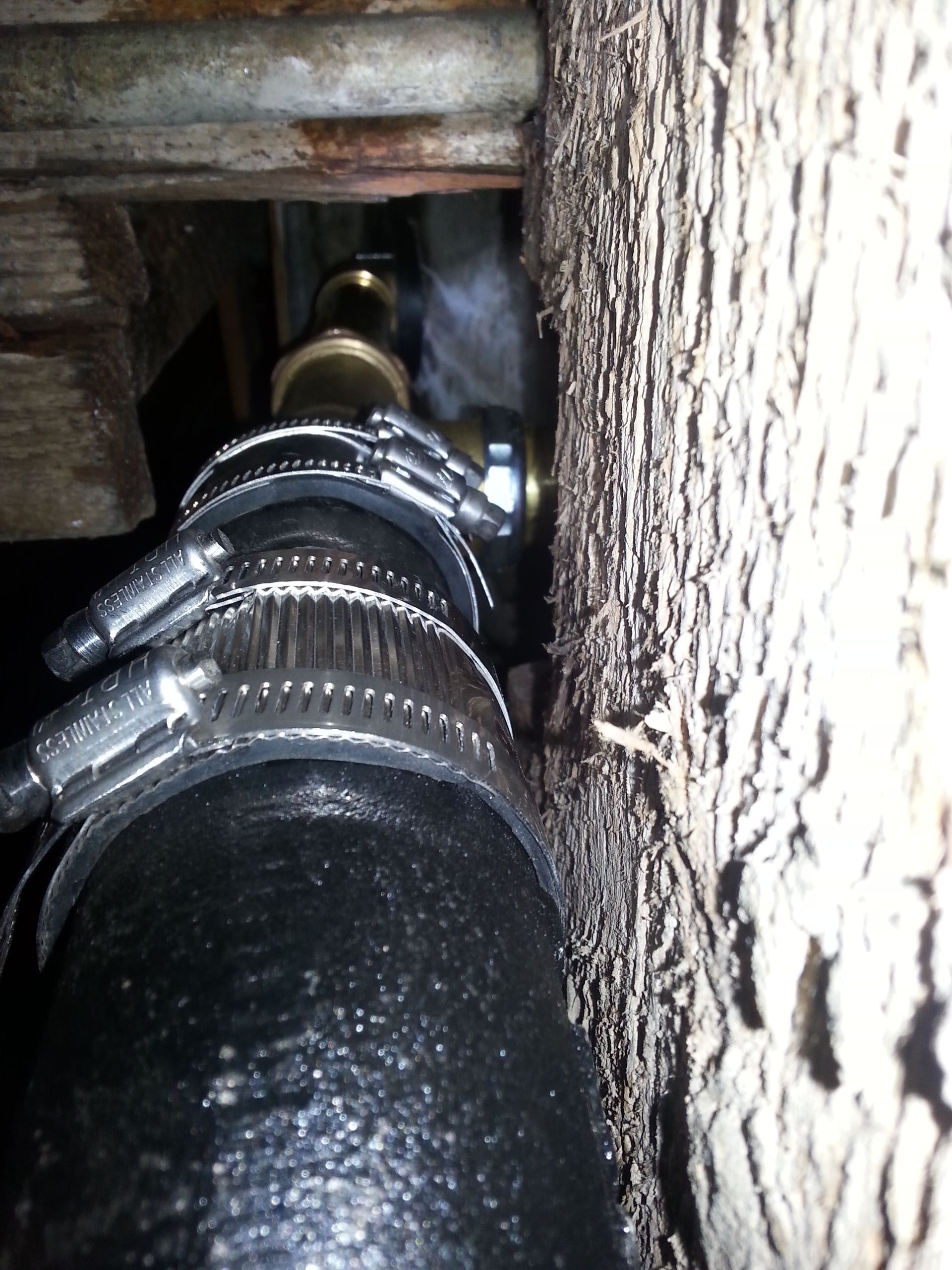 Black pipe connected to brass fitting with clamps against wooden structure.
