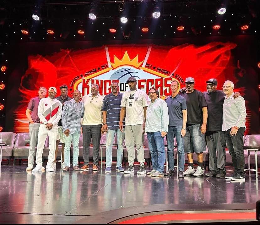 Group of men standing on a stage in front of a red “Kings of... ” backdrop, with bright lights overhead