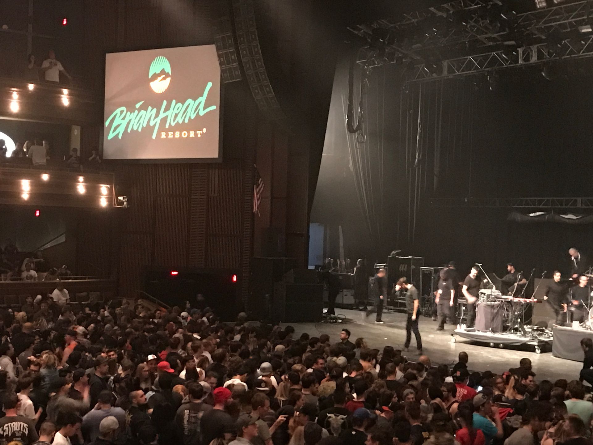 Crowded concert venue with a lit stage, band performing, and a large screen showing the event logo