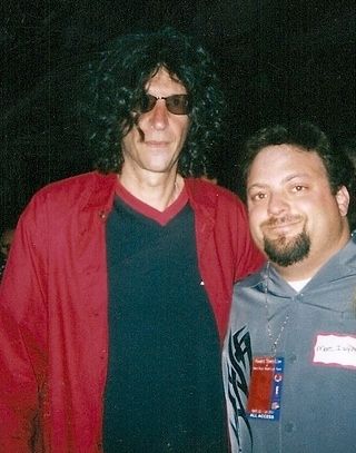 Two people posing together, one in a red jacket and sunglasses, the other in a gray shirt with a lanyard.