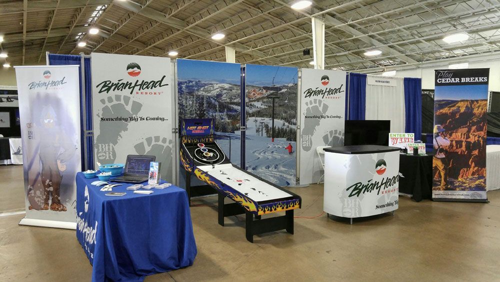 Trade show booth with ski-themed displays, banners, table, and a snowboard bench under a large indoor hall.