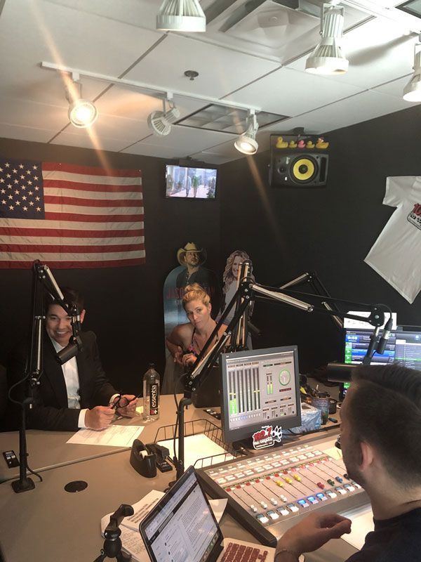 People in a radio studio with microphones, mixing board, and an American flag on the wall.