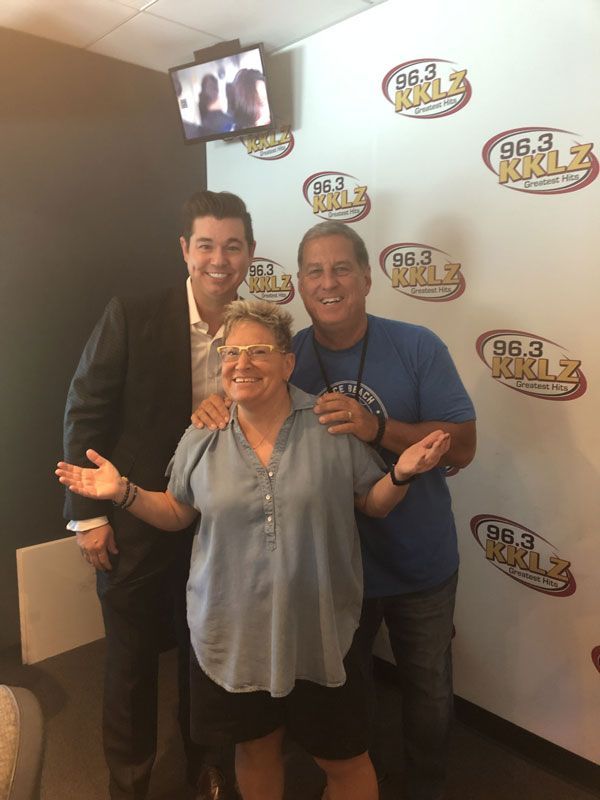 Three people pose smiling in a radio station studio with 98.3 KKLZ logos on the wall.